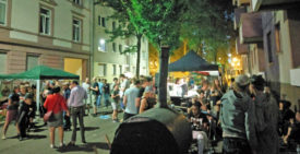 Totale über das Straßenfest. Es ist bereits dämmrig, die Straßenbeleuchtung ist an. Die Besucher scharen sich um Stände oder schauen zur Bühne, die Straße ist voll. Im Vordergrund der mobile Pizzaofen von ZappiZappi