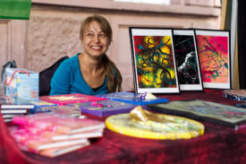 Frau Fenster hinter ihrem Präsentationsstand. Sie lächelt, schaut eine Person außerhalb des Bilds an. vor ihr auf dem Tisch Bilder in Rahmen und auf Leinwand, teils plastisch ausgearbeitet