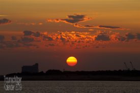 Sonnenuntergang. Eine tiefstehende Sonne stellt sich schon in geld und orange dar, beleuchtet von hinten/unten noch ein paar kleine Schönwetterwolken. Als Silhouette am Horizont sind bauten, mglw. Industriegelände zu erkennen.