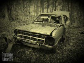 Foto nachbearbeitet auf Sepia-Effekt. Gegenstand ist ein Autowrack. Ein mindestens "Youngtimer", wahrscheinlich inzwischen schon Oldtimer. Auf dem verbeulten Kühlergrill ist gerade noch "Opel" zu erkennen, wahrscheinlich ein Kadett C. Die Scheinwerfer fehlen, ebenso die Windschutzscheibe und andere Fenster. Motorhaube, Kühlergrill und Fahrertür sind verbeult und hängen teils nur lose in den Scharnieren. Die Kofferraumklappe steht offen. Dieses Autowrack steht etwas rätselhaft mitten im Wald, fernab von Straßen und Wegen einfach auf Laub, und Laub liegt auch auf der Motorhaube.