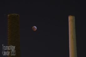 Nachtfoto: Zwei Schornsteine eines Heizkraftwerks, vom städtichen Lichtkonzept leicht angestrahlt. Zwischen den Schornsteinen nicht ganz mittig ein Vollmond in dunkelroter Färbung, vom Blutmond oder ener (partiellen) Mondfinsternis. Einige Betrachter sagten spontan: "Mond-Pong!"